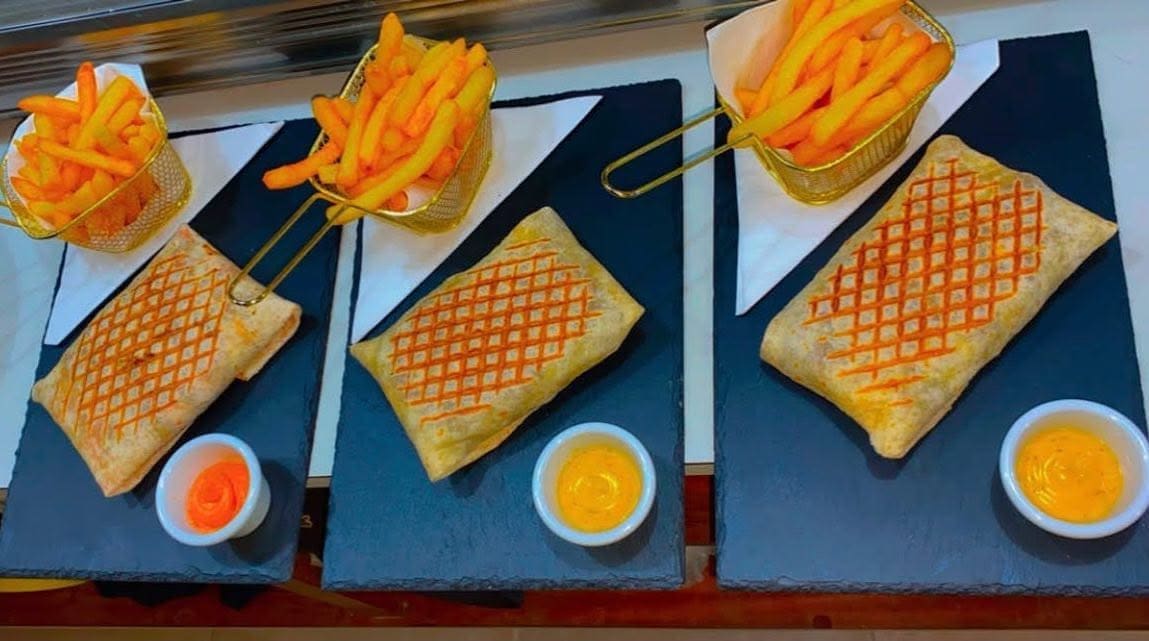 Tres french tacos a la plancha sobre pizarra con patatas fritas y salsas